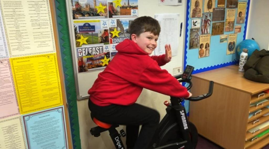 A boy wearing his red school jumper, sat on an exercise bike in a class room. He is smiling and waving. 