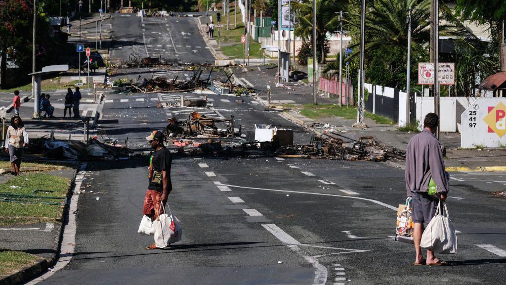 People walk past roadblock barricades set up by rioters near a supermarket, which just reopened to allow people to purchase groceries and food, in Noumea, France's Pacific territory of New Caledonia, on May 18, 2024