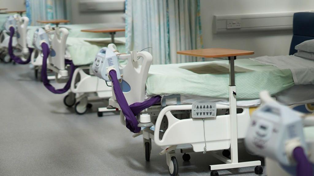 A stock image of a row of inpatient beds at a hospital. They are empty with a swivel table over the bed and medical equipment attached to the plastic frames. Each bed is separated with a drawn-back curtain.