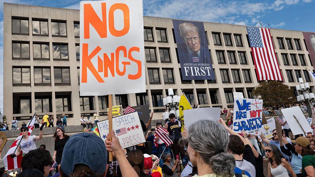 protesters holding up signs at No Kings protest in DC as banner of Trump hanging from government building looms above
