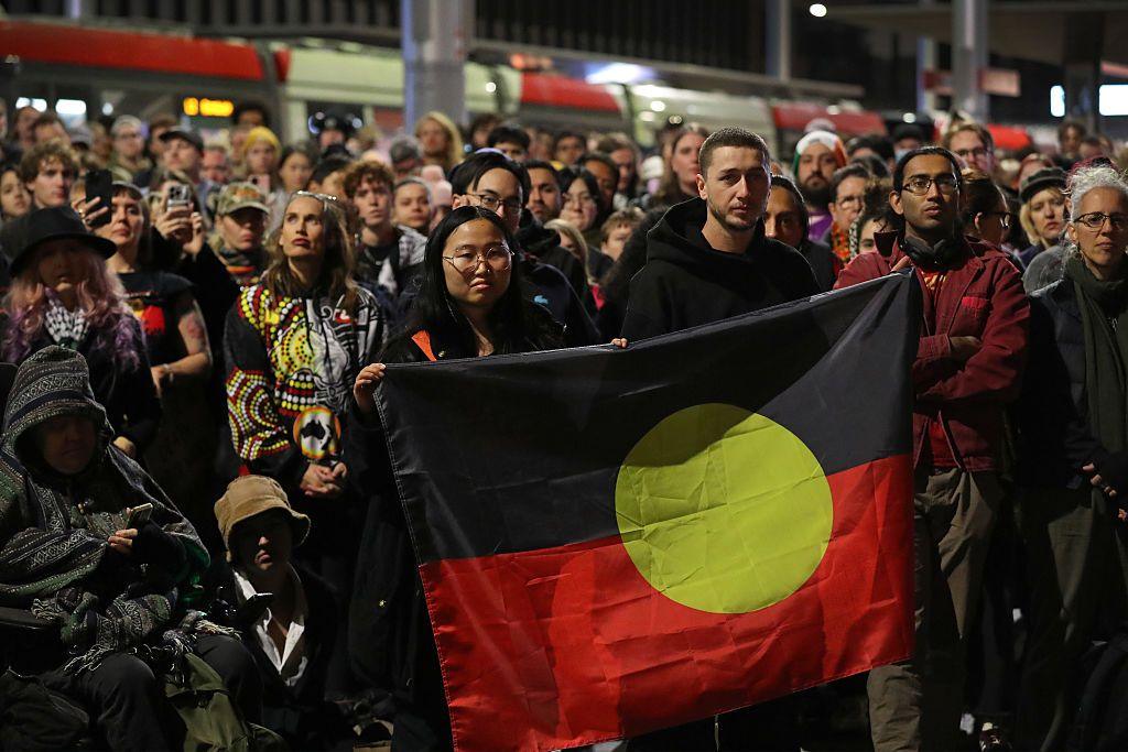 Large group of standing protestors, with several people in the front holding up an Aboriginal flag.