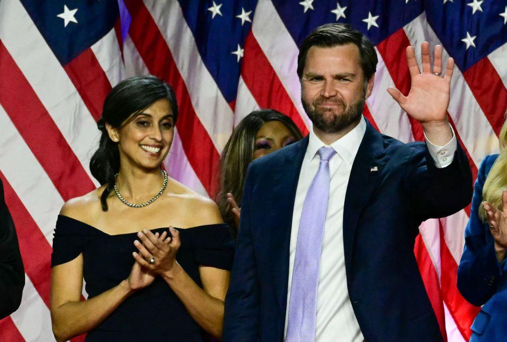 Usha Vance stands with her hands clasped in off-the-shoulder black dress while JD Vance stands next to her in suit and lavender tie, waving his left hand