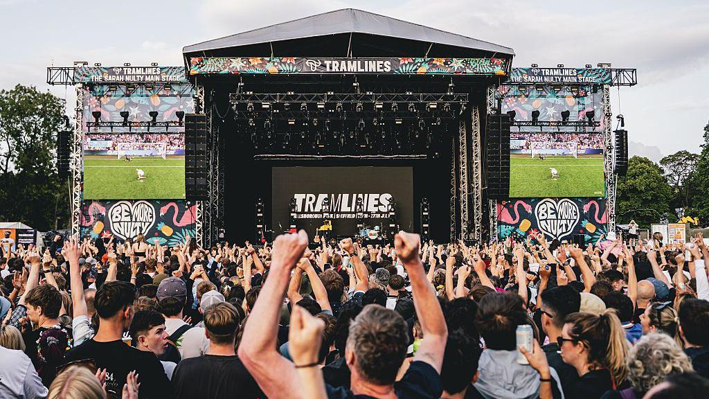 A football game is shown on two screens beside a festival stage, as an onlooking crowd cheer.