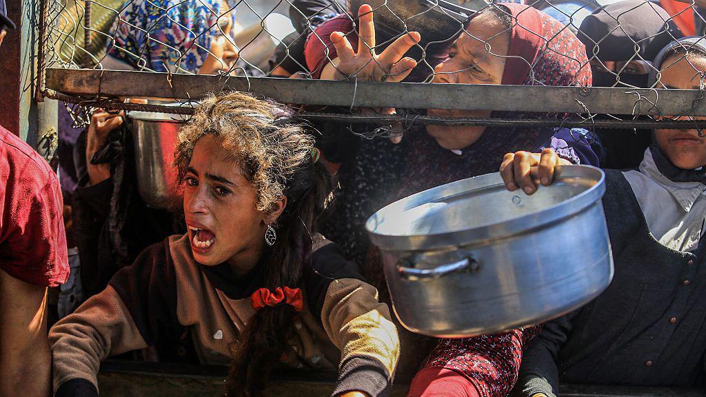 Women and girls hold out pots, one girl crying out, as they press against a fence at an aid distribution point