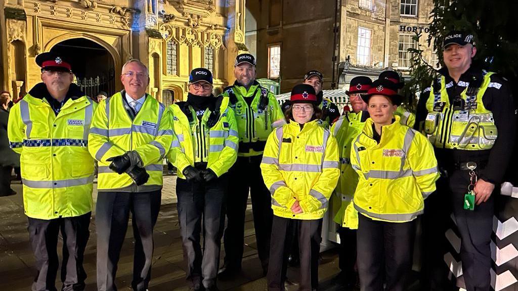 A group of police officers, including Gloucestershire Police and Crime Commissioner Chris Nelson, stand wrapped in hi-vis coats and warm uniform while standing outside a church in Cirencester's Market Place, next to a Christmas tree.