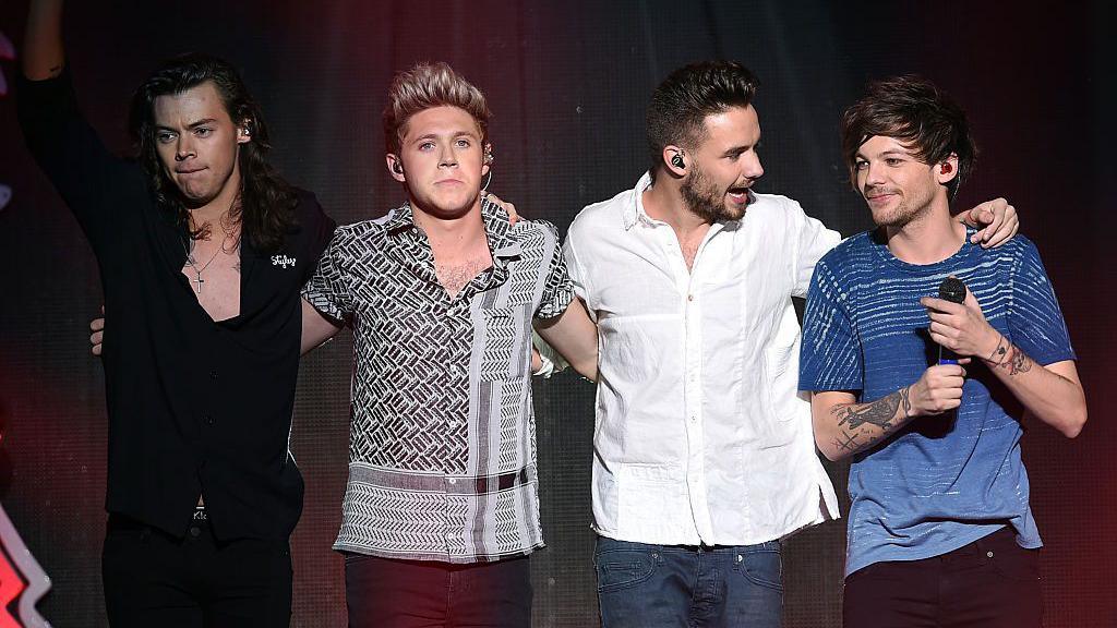 Harry Styles, Niall Horan, Liam Payne and Louis Tomlinson standing in a row. Harry is wearing a black suit and has long brown hair. Niall has blonde short hair and a black patterned shirt. Liam is wearing jeans and a white shirt. Louis has brown short hair and is wearing a blue t shirt, holding a microphone