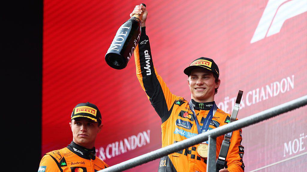 Oscar Piastri holds a bottle of champagne to the crowd after winning at Spa on the podium, with Lando Norris looking on dejected in the background.