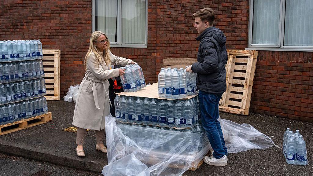 A woman and young adult help to move bottled water supplies from a crate.
