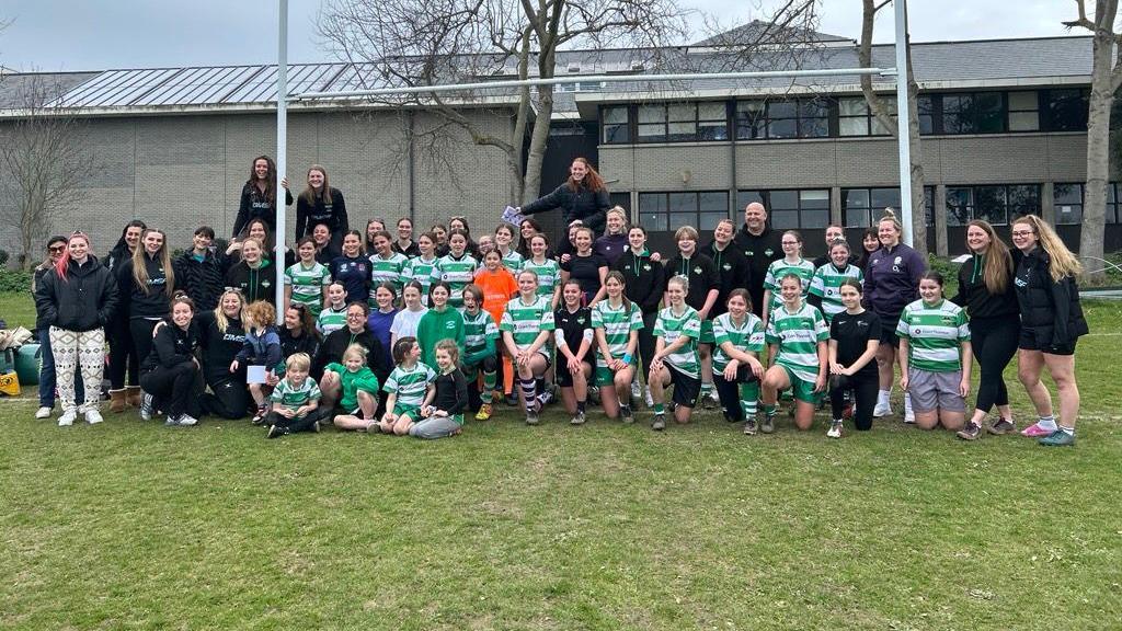 Dozens of girls who play rugby in Guernsey line-up under a set of rugby posts to pose for a team photo. They are joined by some of their coaches and England stars Marlie Packer and Rosie Galligan. Some of the girls are wearing the green and white hooped playing tops worn by Guernsey's team.