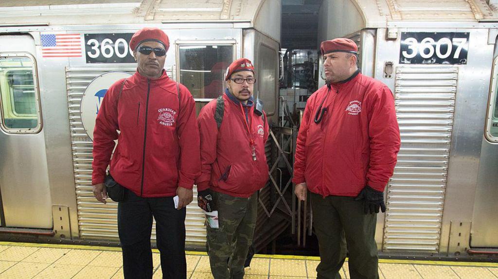 Three Guardian Angels security guards standing in front of a subway train in New York. They are wearing the red jackets the organisation is well known for. The tallest guard is wearing black sunglasses, the one in the middle has normal glasses, and all three are wearing red berets.