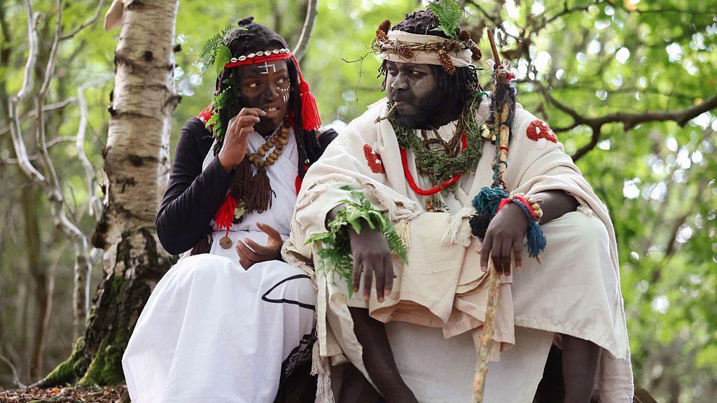A man and a woman dressed in white/cream robes in a woodland setting. They have painted faces and head-dresses. They have leaves in their hair. 
