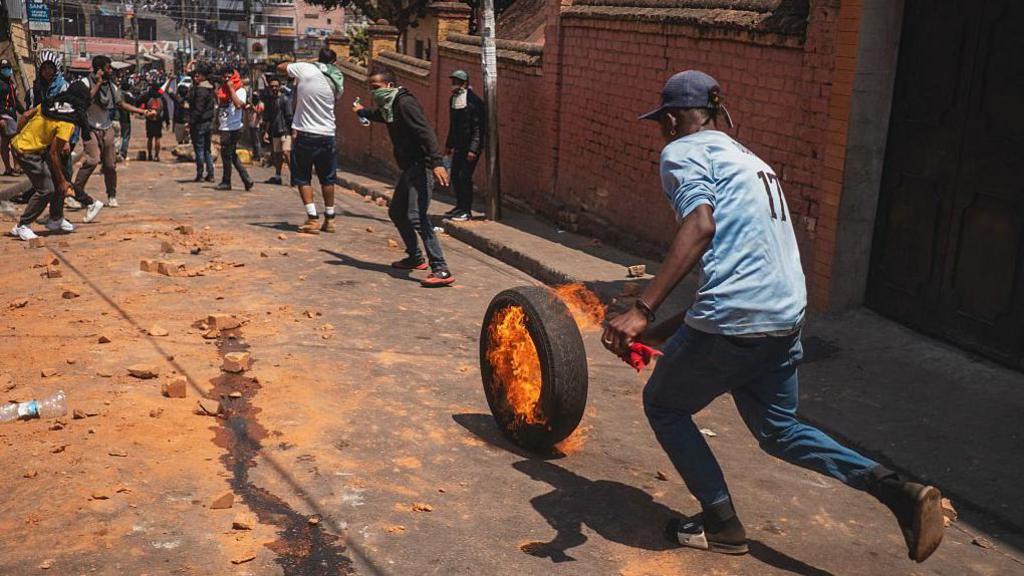 A man runs behind a tyre that is on fire and rolling along a road.