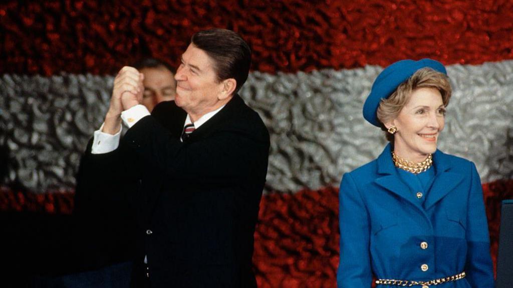 Ronald Regan and his wife Nancy stand in front of the American flag. Both are smiling. President Reagan clasps his hands in thanks to the crowd