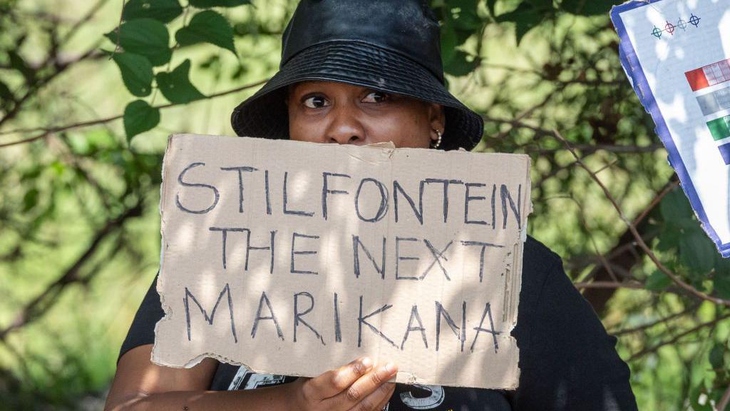 A woman in a black sun hat stands with a handmade cardboard placard  that says "Stilfontein - the next Marikana"