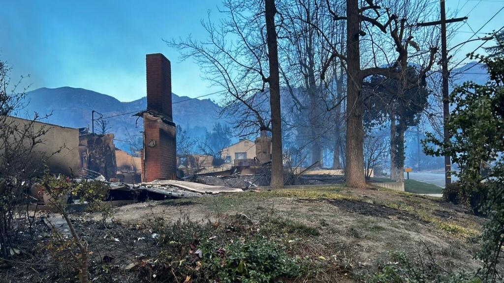 The remains of the house after the fire, with the chimney still standing and blackened rubble around it. There are burnt trees to the side and mountains in the background, with smoke still visible in the air.