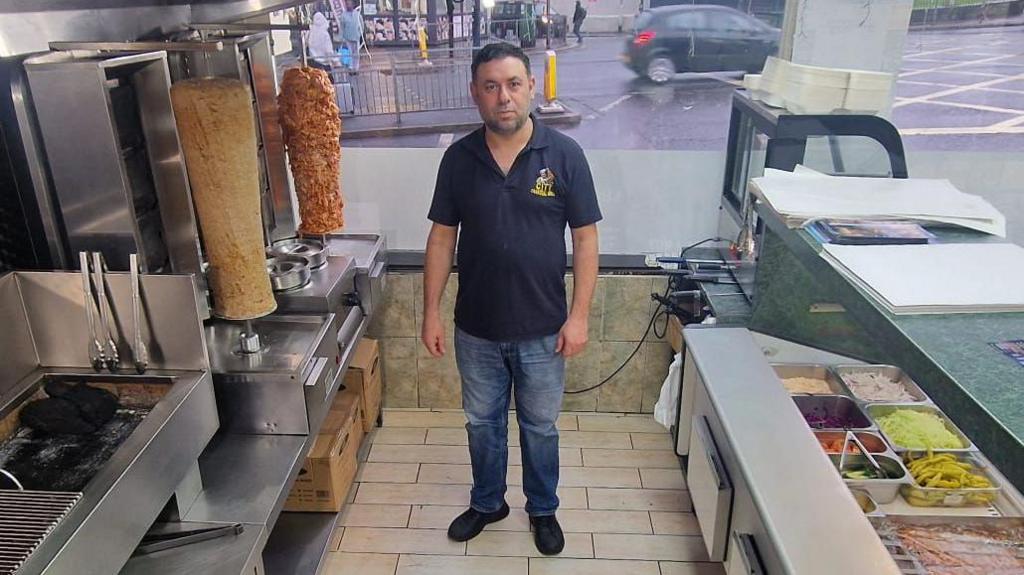 A man in a blue short sleeve top and jeans stands in a kebab shop behind the counter with a street in the background.