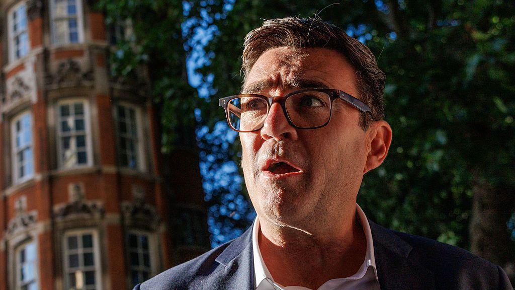 Andy Burnham, wearing a suit, white collared shirt and glasses. A red-brick building and a tree are in the background.