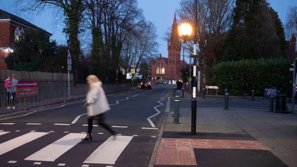 A general view of a road in Birmingham on a dark night. A blurry figure walks across a zebra crossing. There is a building in the background and we can see a car driving along with its lights on.