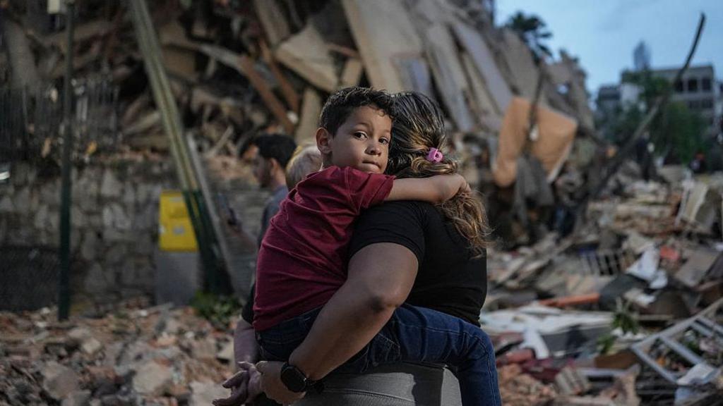 A boy is held by his mother in front of a mountain of rubble in Venezuela. He is looking directly at the camera, has short brown hair and wears a red t-shirt and blue trousers. He is being carried by his mother, who is looking away towards the rubble behind them. She has curly dyed blonde hair and wears a black t-shirt.