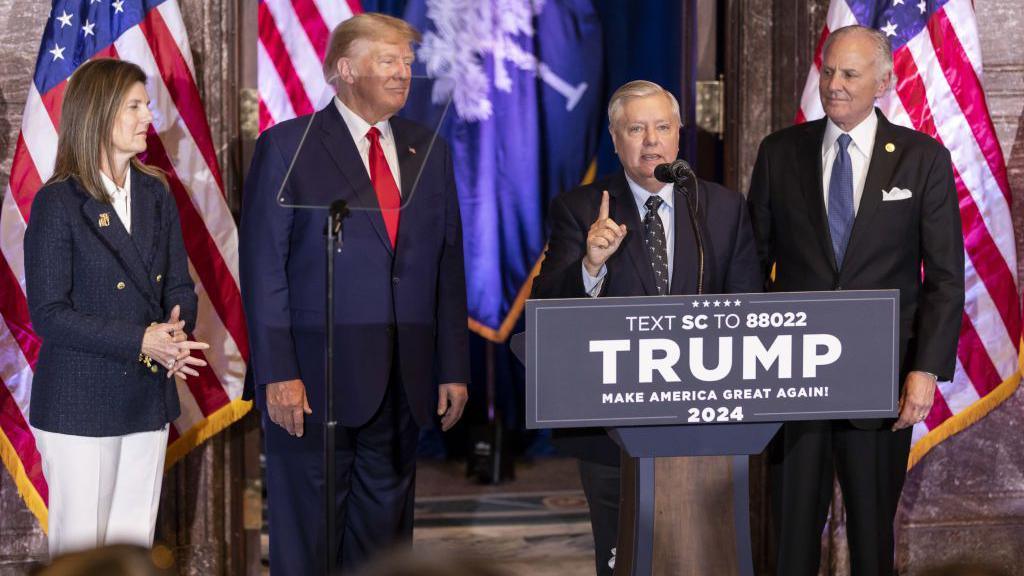 Lindsay Graham speaking at a campaign event for Donald Trump. He is standing behind a lectern marked "Trump" and is standing beside Trump.