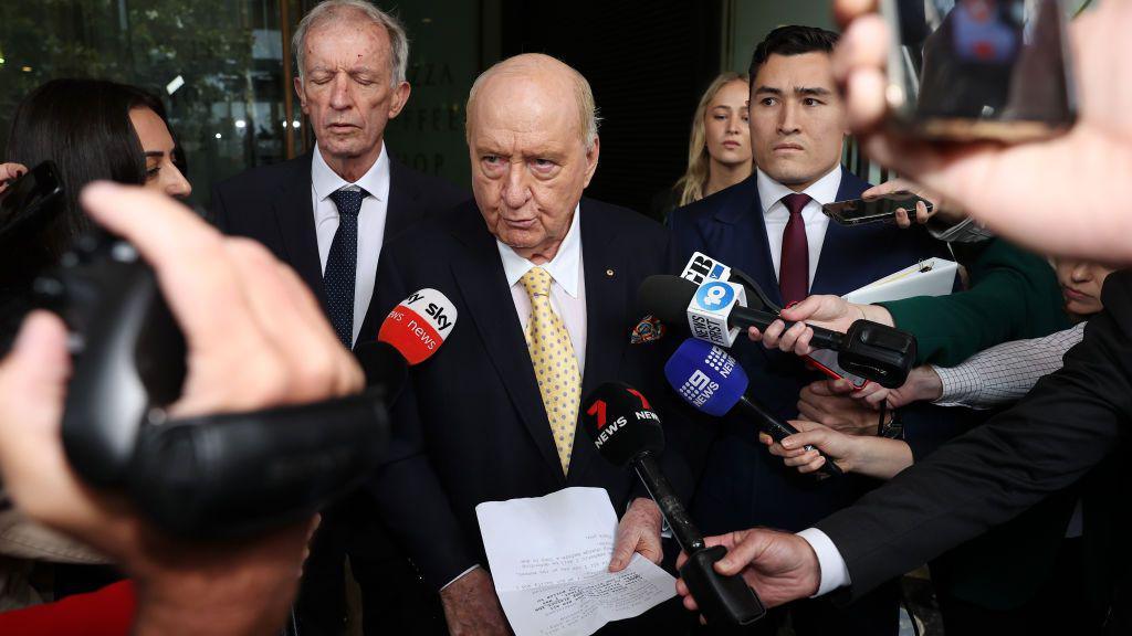 Alan Jones flanked by his lawyers speaks into news microphones while holding a piece of paper.