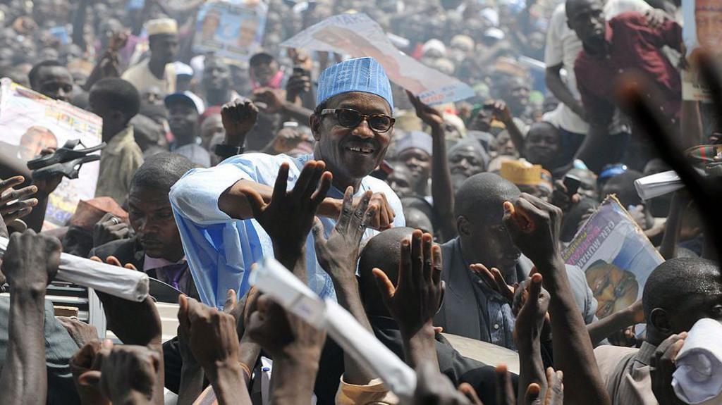 At a packed rally, supporters clamour to touch Buhari, who stands above the crowd, smiling. He wears a blue top and blue hat.