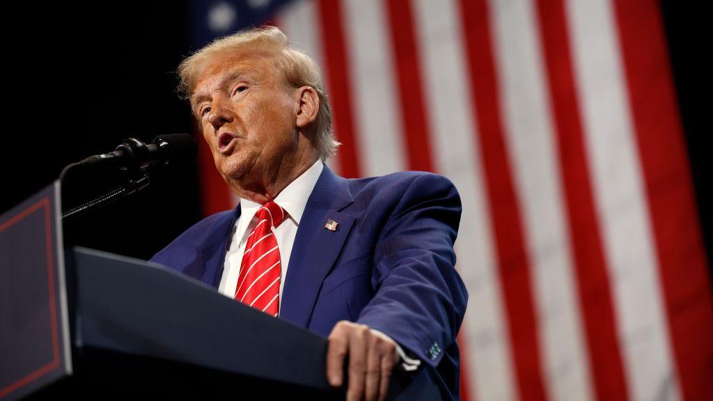 Donald Trump stands at a podium wearing a blue suit and red striped tie with an American flag in the background