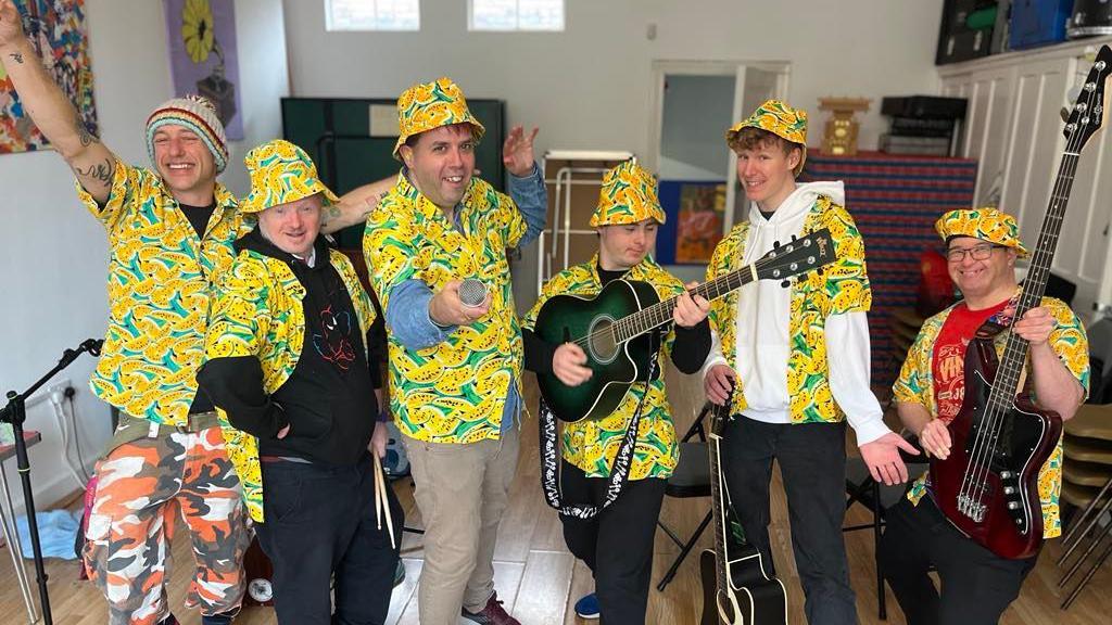 Six performers, all dressed in colourful shirts and hats, smiling and looking towards the camera. Two are holding guitars, one a microphone, standing in a room with instruments around them. 
