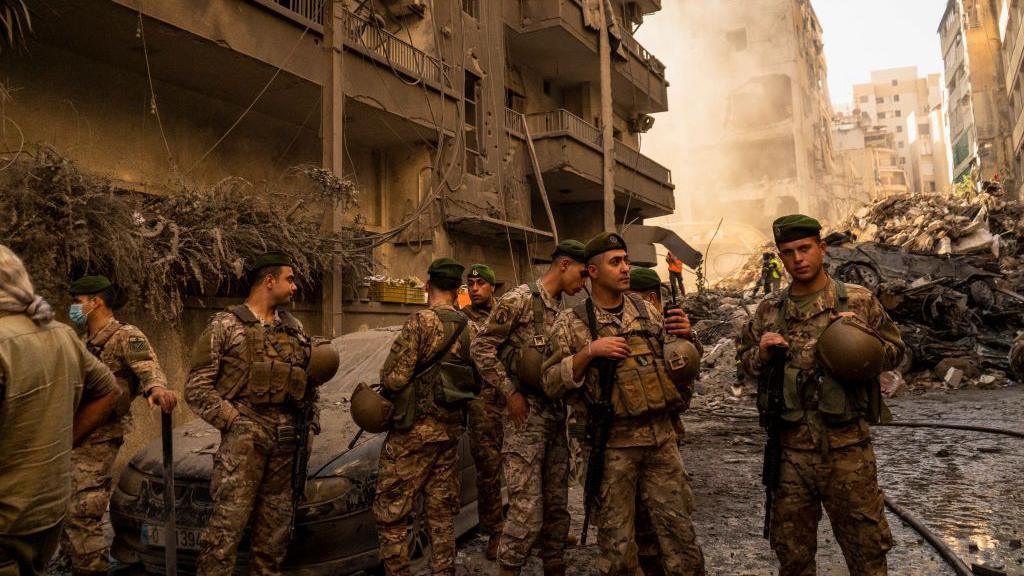 Lebanese Army soldiers stand amidst rubble and damaged buildings in central Beirut, surveying the aftermath of an Israeli airstrike, with smoke and destruction visible in the background.