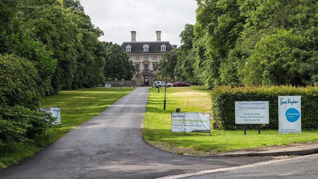 Street view of the Thorpe Hall hospice- a characterful yellow building with a brown stone roof- with a lawned driveway leading up to it