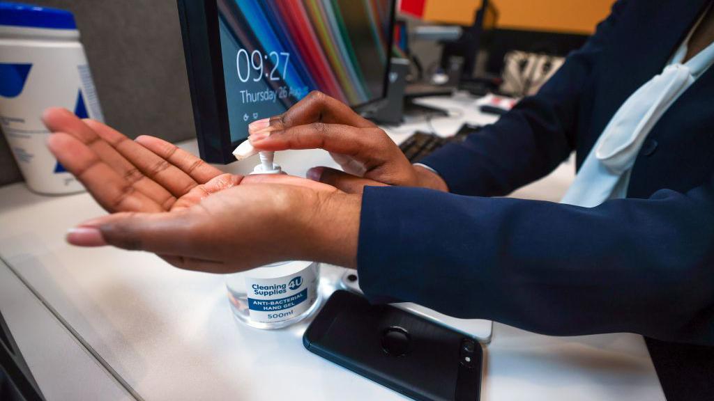 A stock image of a person applying hand sanitiser at a work desk. A computer screen can be seen in the background.