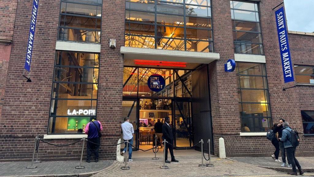 The front entrance of a food hall located in an old, brick warehouse building. People can be seen standing around outside the entrance. There are blue vertical banners on the building that say "Saint Paul's Market". The inside of the market has been lit up and stalls, tables and chairs are visible.
