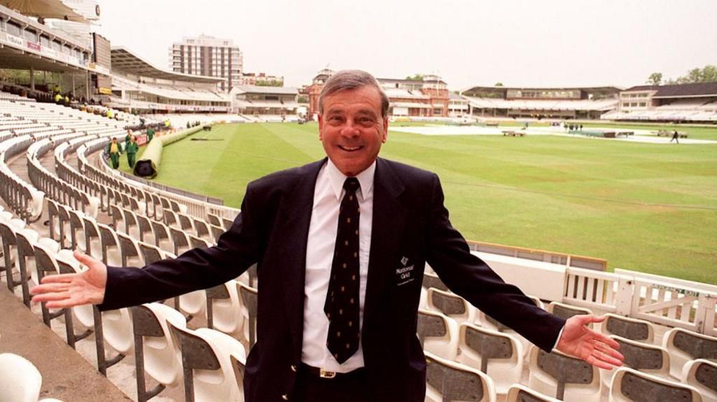 Dickie Bird smiles with his hands outstretched, with a cricket ground behind him.