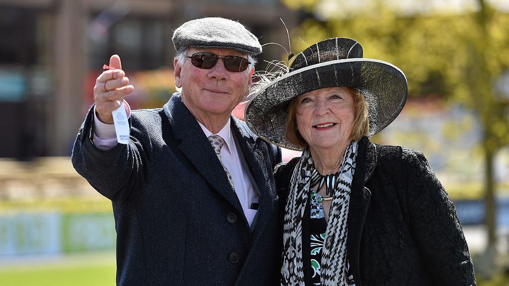 A man and a woman look, smiling, at the camera. The man is wearing a jacket, a white shirt, a tie, a hat, and sunglasses. The woman is wearing a jacket, a scarf, and a hat. The weather looks sunny and, in the distance, there is a tree.