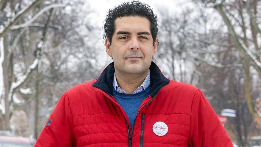 Vince Gasparro, he is looking at the camera, he has black hair and is wearing a blue jumper and shirt with a red jacket.