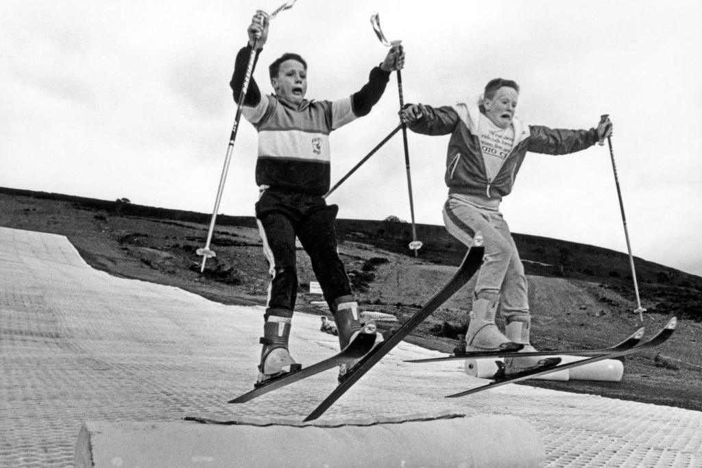 Children practising their ski-jumps on their self-made jumps on an artificial ski slope. The picture is in black and white and the two young boys are in mid-air. A featureless hill is behind them