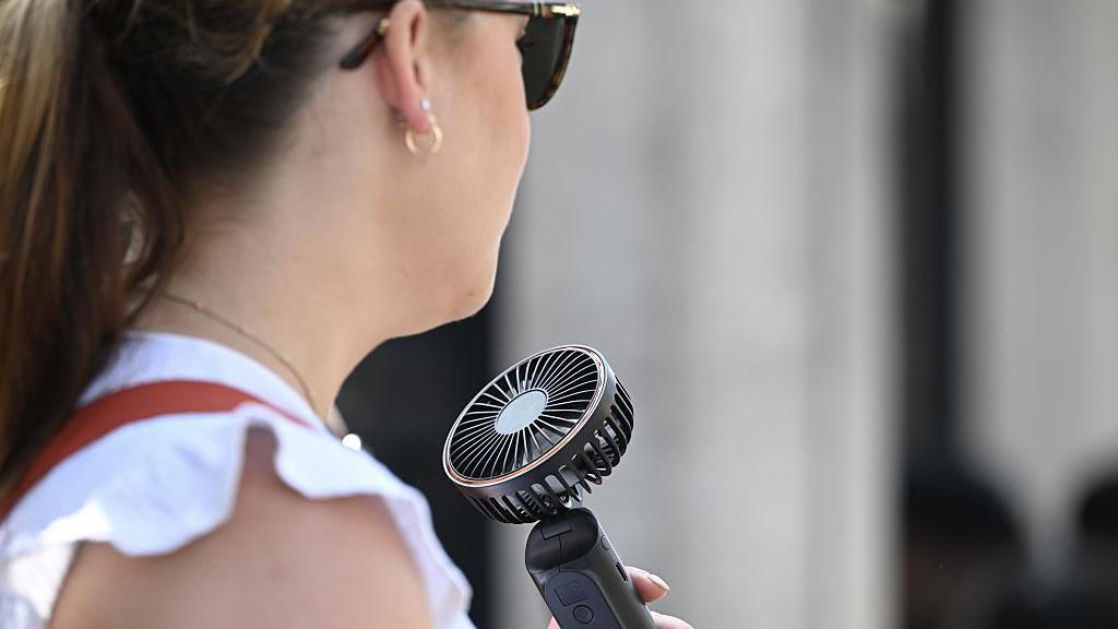 A woman with dark hair and wearing sunglasses and a white blouse, standing outside and holding a handheld fan up to her face.