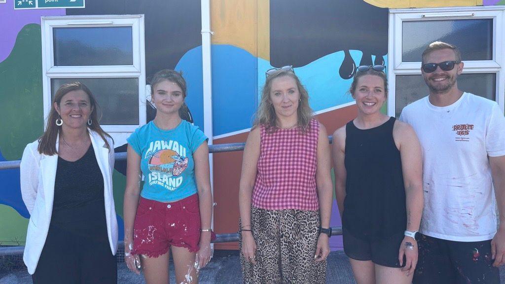Four women and one man standing in front of a painted building. The building has many bright colours and different shapes. The people are all standing next to each other smiling.