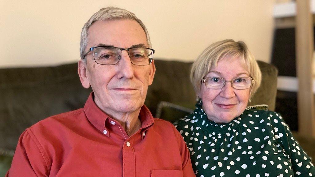 Chris ac Alyson Austin yn eu stafell byw yn edrych yn syth at y camera. Maent wedi gwisgo'n smart.