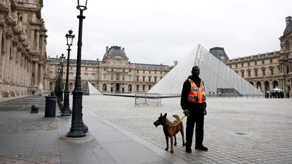 The Louvre is seen from the outside, where the pyramid is, with a security guard standing with a dog