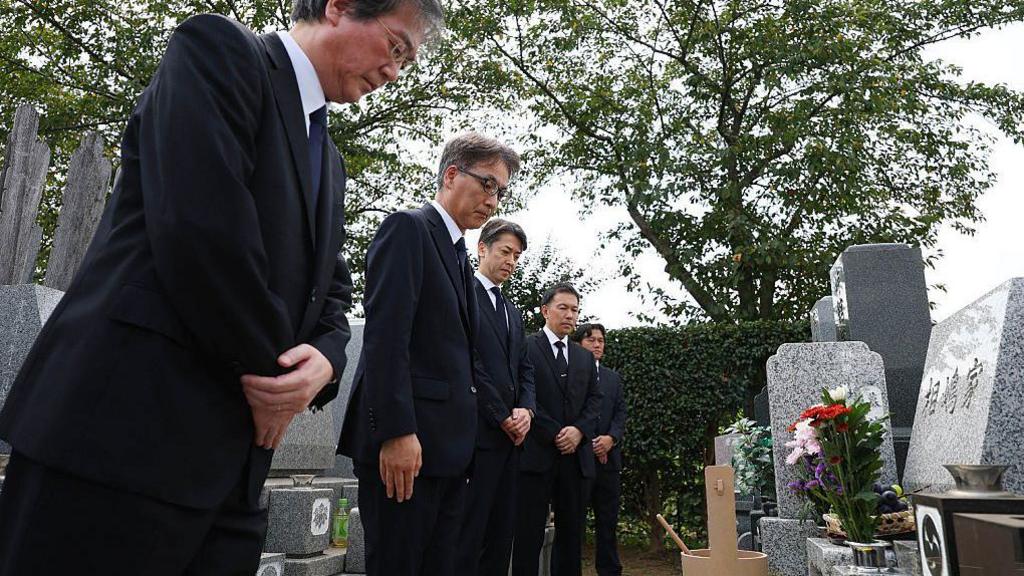 Five men wearing dark suits bow before a grave in Japan. The grave is made of grey marble, and has flowers in a vase in front of it