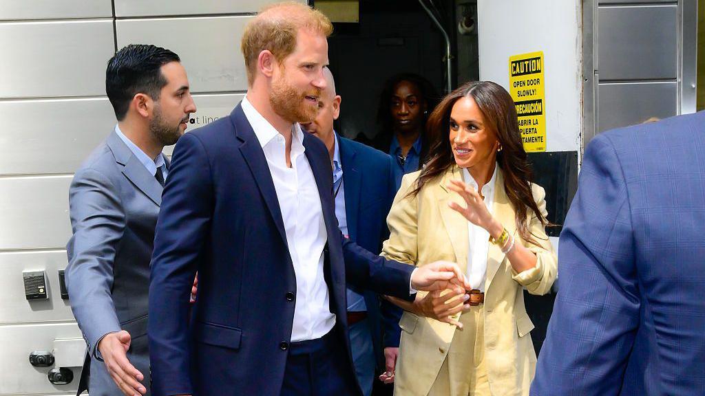 The Duke and Duchess of Sussex walk out of a door. The duchess is waving and smiling. Prince Harry is wearing a white shirt and a black blazer and trousers. Meghan is wearing a white shirt and a beige blazer and trousers. Prince Harry has ginger hair, a ginger beard, and blue eyes. Meghan has long brown hair and brown eyes.