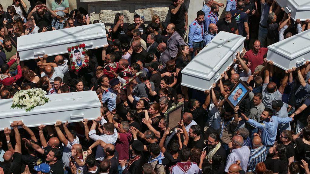 A birds eye view shows five white coffins being carried over people's heads with a large crowd of mourners gathered around them