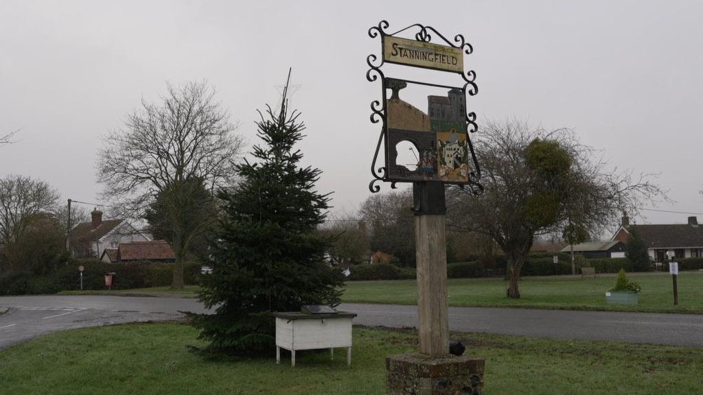 A view of a village square with a tall sign that reads "Stanningfield". A tree next to it can be seen while homes are in the background along with a village green. 