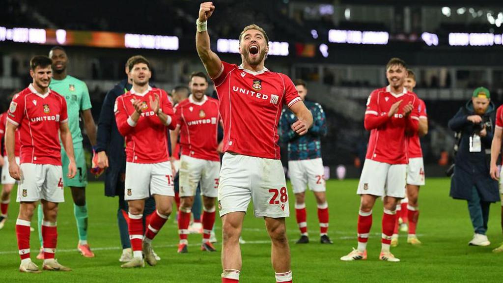 Wrexham striker Sam Smith celebrates with his fist raised, while his team-mates stand behind and applaud