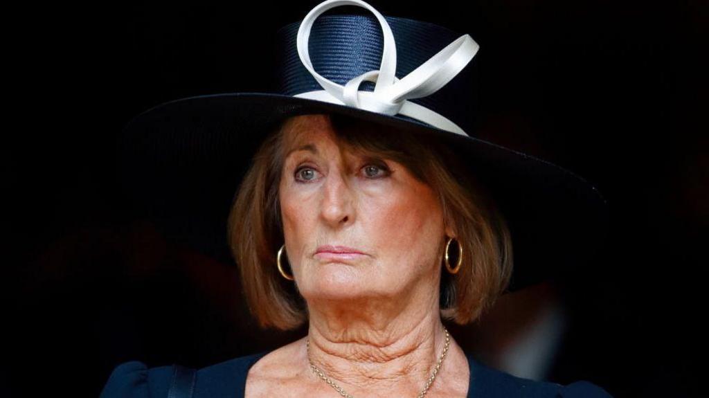 Annabel Goldsmith wearing a dark blue formal hat and dress. The hat has a cream-coloured bow. She is wearing gold hoop earrings.