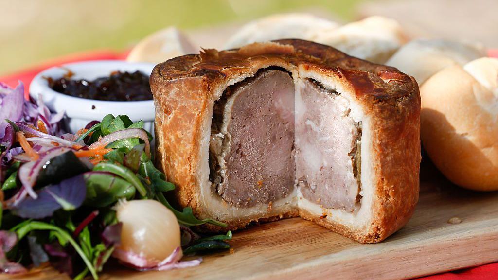 A photograph of a Melton Mowbray pork pie, with a slice missing, surrounded by salad to the left and a piece of bread to the right. 