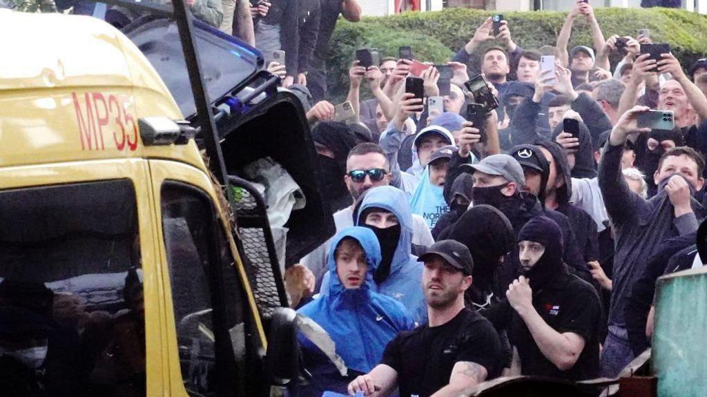 A crowd of people, some with their faces covered, standing in front of a police van. Some appear to be throwing a wheelie bin while many others are holding up their phones to film the scene