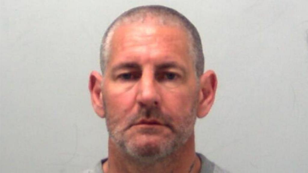 A custody photo of Mitchell Rudkin. He is wearing a grey jumper and has a shaved head.