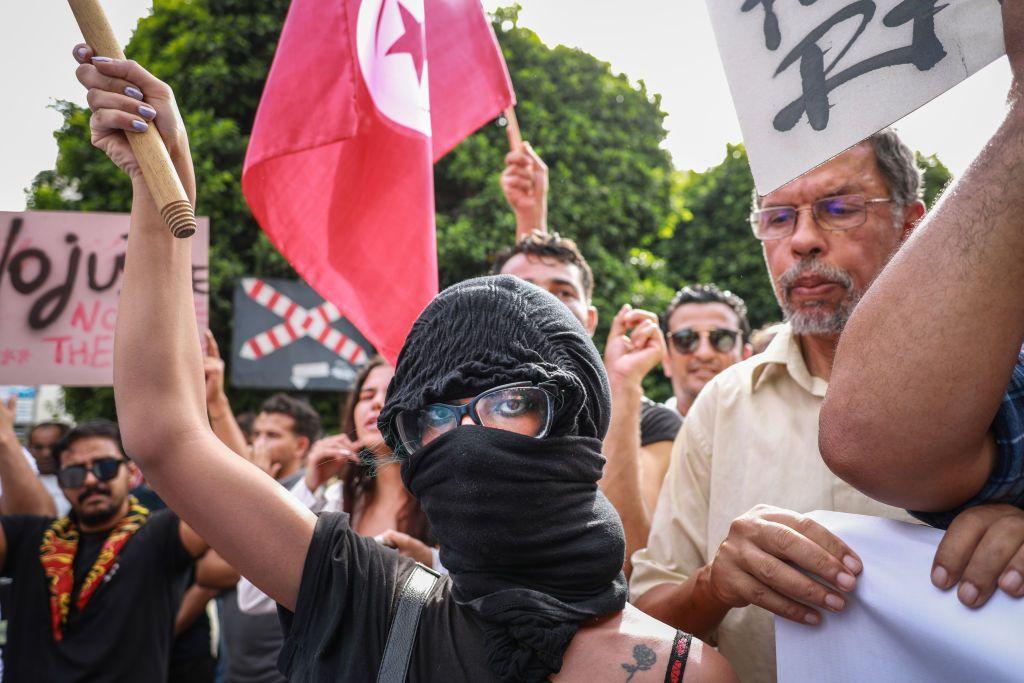 A young female demonstrator with her face covered by a bandanna raises her arm as others wave the national flag of Tunisia during a demonstration on September 22, 2024